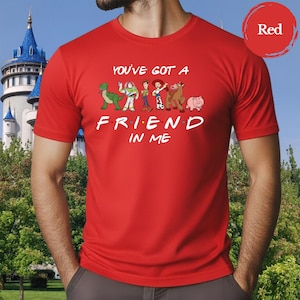 May include: A red t-shirt featuring the phrase "YOU'VE GOT A FRIEND IN ME" with cartoon character graphics. The design includes a green dinosaur, a space ranger, a cowboy, a cowgirl, a horse, and a pig.