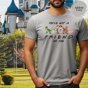 May include: Athletic grey t-shirt featuring the phrase "You've Got a Friend in Me" with cartoon character graphics. The design includes a green dinosaur, Buzz Lightyear, Woody, Jessie, Bullseye, and Hamm. The shirt is made of a soft material.