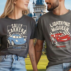 May include: Two charcoal gray t-shirts featuring characters from the movie Cars. One shirt displays "SALLY CARRERA" with a blue car graphic and the text "I'M LIGHTNING!". The other shirt shows "LIGHTNING MCQUEEN" with a red car graphic and the text "I'M LIGHTNING!"