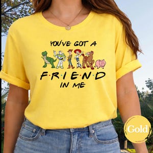 May include: Yellow t-shirt featuring the phrase "YOU'VE GOT A FRIEND IN ME" with cartoon characters. The design includes a green dinosaur, Buzz Lightyear, Woody, Jessie, a horse, and a pig. The shirt is a bright yellow color.
