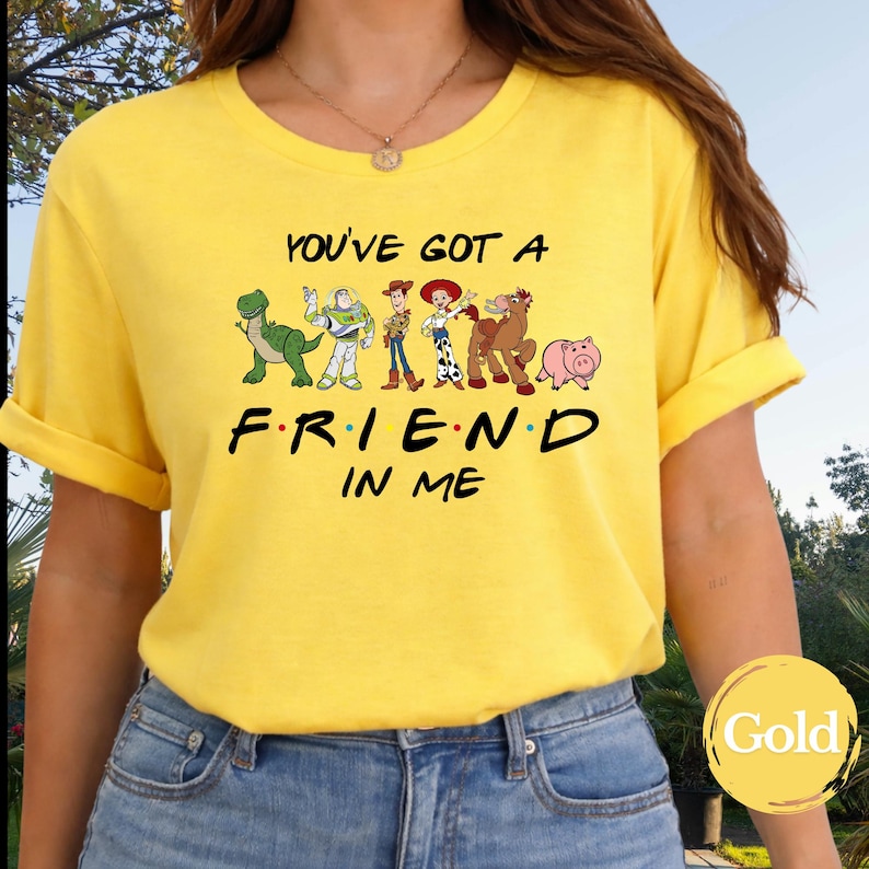 May include: Yellow t-shirt featuring the phrase "YOU'VE GOT A FRIEND IN ME" with cartoon characters. The design includes a green dinosaur, Buzz Lightyear, Woody, Jessie, a horse, and a pig. The shirt is a bright yellow color.