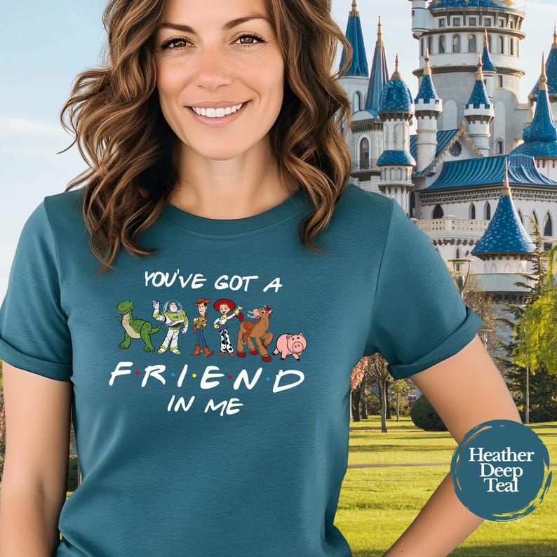 May include: Teal t-shirt featuring the phrase "You've Got a Friend in Me" with cartoon characters. The shirt has a crew neck and short sleeves. The background shows a castle and a blue circle with the words "Heather Deep Teal".