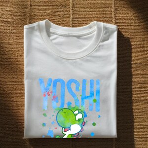 May include: White t-shirt with the word "YOSHI" in blue, and a green cartoon character. The design includes colourful paint splatters. The shirt is folded on a textured surface.