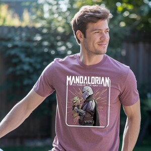 May include: Maroon t-shirt featuring a graphic of The Mandalorian holding Grogu, with the text "MANDALORIAN - THIS IS THE WAY" in white. The design is framed in a white rectangle. The shirt is short-sleeved.