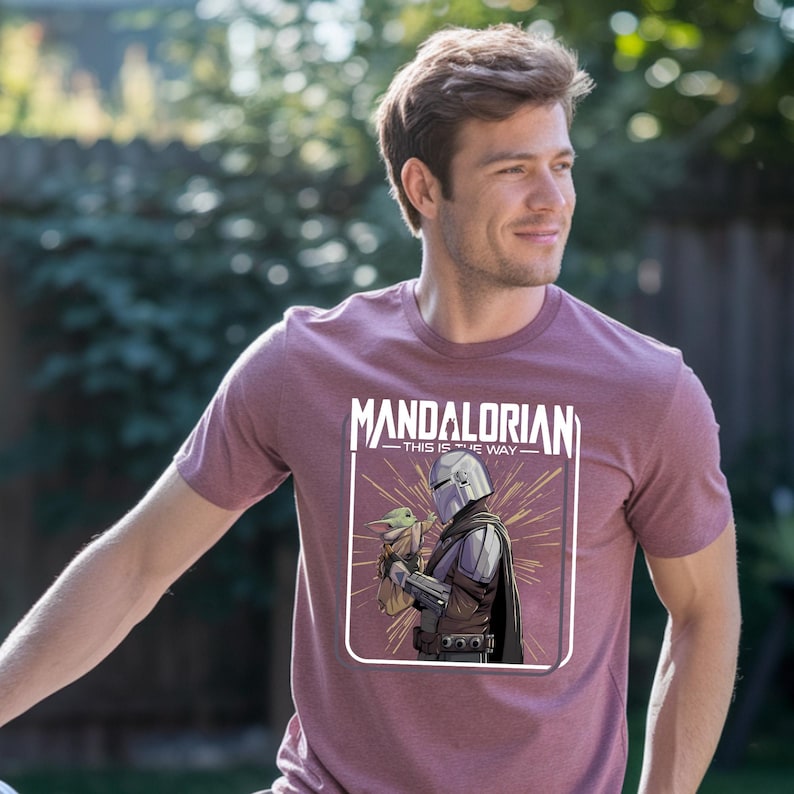May include: Maroon t-shirt featuring a graphic of The Mandalorian holding Grogu, with the text "MANDALORIAN - THIS IS THE WAY" in white. The design is framed in a white rectangle. The shirt is short-sleeved.