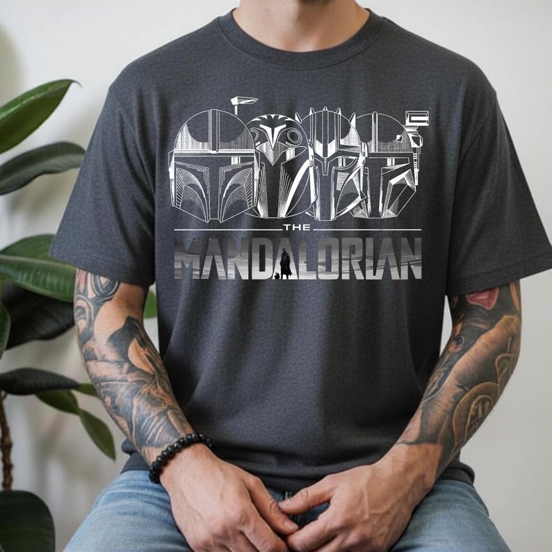 May include: Dark gray t-shirt featuring a white graphic of Mandalorian helmets and the text "THE MANDALORIAN". The design includes a small silhouette of a figure. The shirt is short-sleeved and made of a soft material.