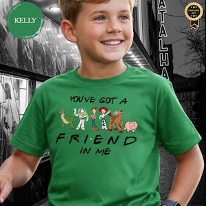May include: A green t-shirt featuring the phrase "You've Got A Friend In Me" with cartoon character graphics. The shirt has a crew neck and short sleeves. The design includes a dinosaur, Buzz Lightyear, Woody, Jessie, a horse, and a pig.