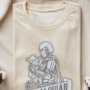 May include: Cream-coloured t-shirt featuring a graphic of The Mandalorian holding Grogu. The design is in shades of grey and white, with the text "The Mandalorian" at the bottom. The shirt has a crew neck.