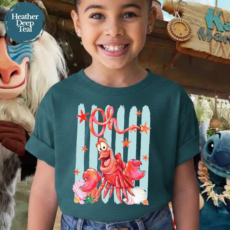 Pu&ograve; includere: Una t-shirt a maniche corte color verde acqua scuro con la grafica di un granchio rosso e la scritta Oh in corsivo sopra. Il design include strisce verticali azzurre, piccole stelle marine arancioni e conchiglie bianche.