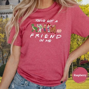May include: Red t-shirt featuring the phrase "You've Got a Friend in Me" with cartoon characters. The design includes a green dinosaur, a space ranger, a cowboy, a horse, and a pig. The shirt is a solid red color.