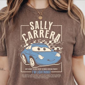 May include: Brown t-shirt featuring a blue cartoon car with the text "SALLY CARRERA" above it. The text "I'M LIGHTNING!" is below the car. The shirt has a vintage, distressed look.