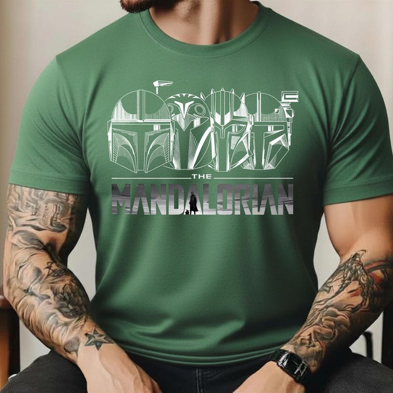 May include: Green t-shirt featuring a white graphic of Mandalorian helmets and the text "THE MANDALORIAN" in silver. The design includes a small silhouette of the Mandalorian. The shirt is made of a soft material.