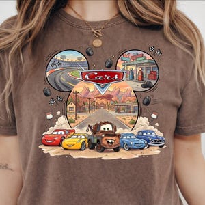 May include: Brown t-shirt featuring a colorful graphic of characters from the animated movie "Cars." The design includes race tracks, a desert landscape, and the word "Cars" in a stylized font. The shirt has a crew neck.