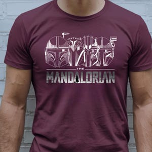 May include: Burgundy t-shirt featuring a white graphic of Mandalorian helmets and the text "THE MANDALORIAN". The design includes detailed helmet illustrations and a stylized font.