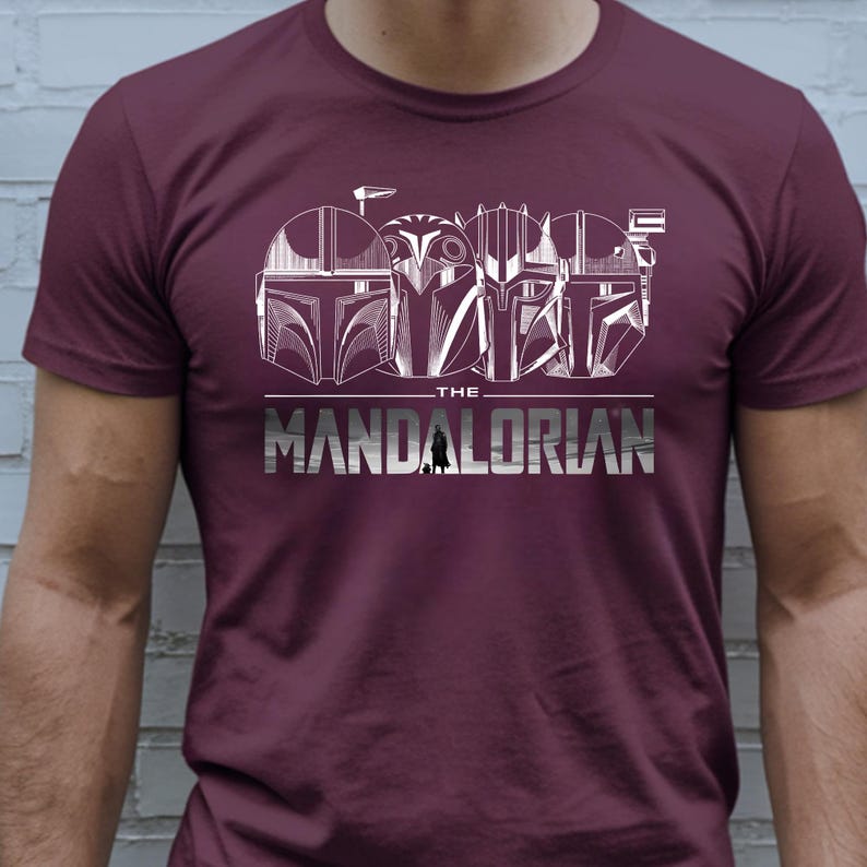 May include: Burgundy t-shirt featuring a white graphic of Mandalorian helmets and the text "THE MANDALORIAN". The design includes detailed helmet illustrations and a stylized font.