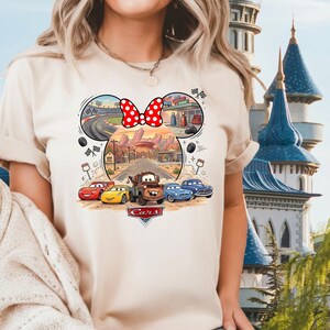 Pode incluir: Uma t-shirt bege de manga curta com uma impress&atilde;o gr&aacute;fica das personagens do filme Carros da Disney Pixar, incluindo Rel&acirc;mpago McQueen e Mate. O design tem a forma de uma cabe&ccedil;a do Mickey Mouse com um la&ccedil;o vermelho de bolinhas brancas.