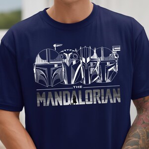 May include: Navy blue t-shirt featuring a white graphic of Mandalorian helmets and the text "THE MANDALORIAN". The design includes a small silhouette of the Mandalorian character.