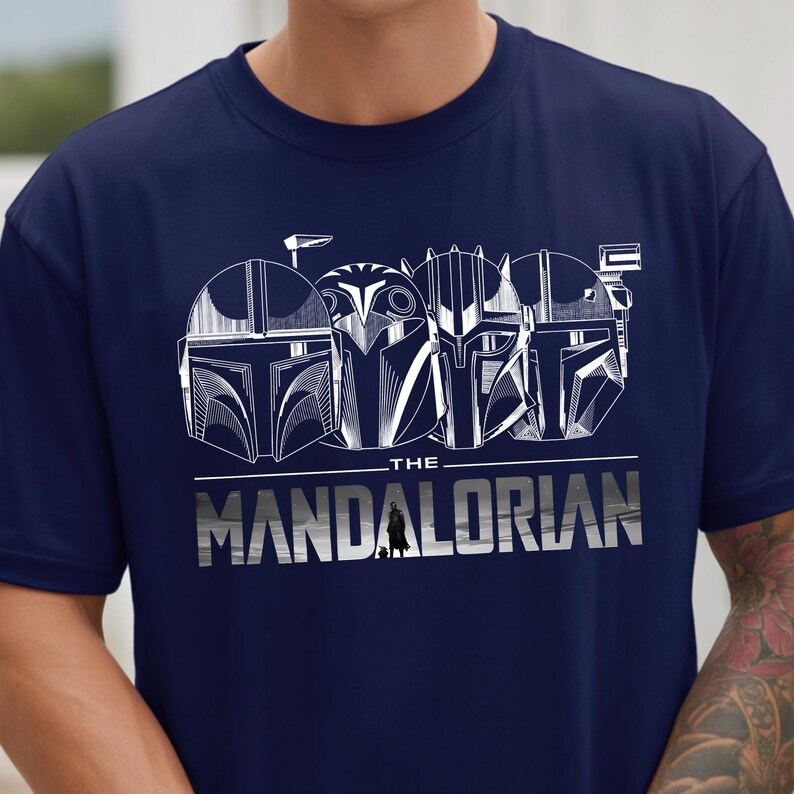 May include: Navy blue t-shirt featuring a white graphic of Mandalorian helmets and the text "THE MANDALORIAN". The design includes a small silhouette of the Mandalorian character.