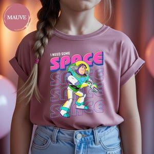 May include: Mauve-colored t-shirt featuring Buzz Lightyear from Toy Story. The graphic includes the text "I NEED SOME SPACE" in a colorful, retro font. Buzz Lightyear is depicted in his signature space suit. The shirt is made of soft material.