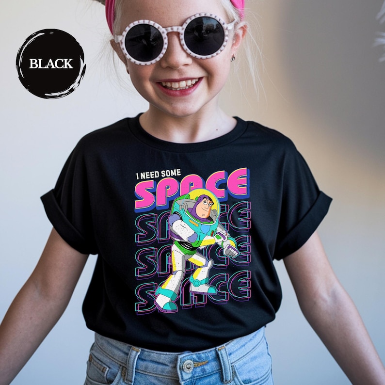 May include: Black t-shirt featuring a colorful graphic of Buzz Lightyear with the text "I NEED SOME SPACE". The design includes the word "SPACE" in pink and blue, with a repeating effect. The shirt is a crew neck style.