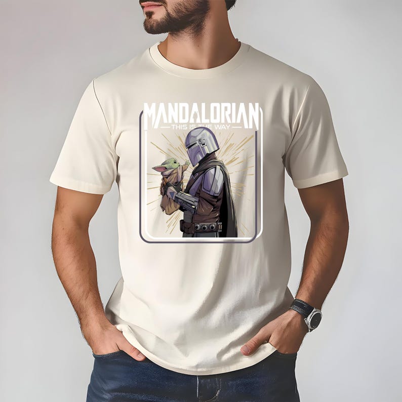 May include: Beige t-shirt featuring a graphic of The Mandalorian holding Grogu, with the text "MANDALORIAN - THIS IS THE WAY" in a white square. The illustration shows the characters in detailed armor and clothing.