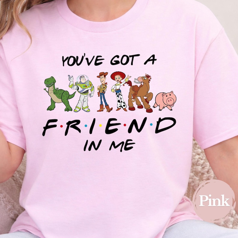 May include: A light pink t-shirt featuring the phrase "YOU'VE GOT A FRIEND IN ME" with cartoon character illustrations. The characters include a green dinosaur, Buzz Lightyear, Woody, Jessie, Bullseye, and Hamm.