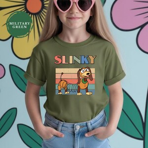 May include: A military green short-sleeve t-shirt featuring a graphic of the Slinky Dog character from Toy Story. The design includes the text SLINKY in colorful letters above the dog, set against a retro-style horizontal striped background.