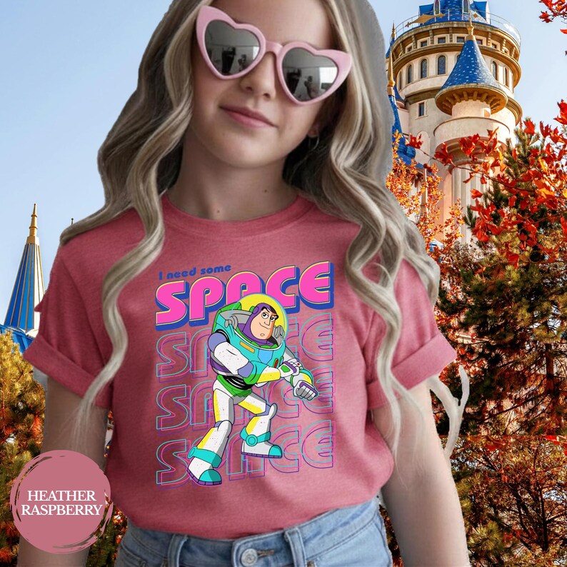 May include: Heather raspberry t-shirt featuring a graphic of Buzz Lightyear with the text "I need some SPACE". The design includes the word "SPACE" in a vibrant blue and pink font, repeated vertically. The shirt is a crew neck.