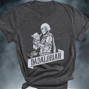 May include: Dark grey t-shirt with a white graphic of a character in armour holding a small creature. The text "The DADALORIAN" is displayed in a white banner below the graphic.