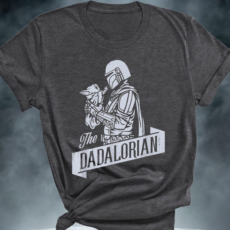 May include: Dark grey t-shirt with a white graphic of a character in armour holding a small creature. The text "The DADALORIAN" is displayed in a white banner below the graphic.
