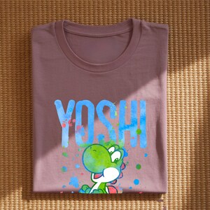 May include: A mauve t-shirt with the word "YOSHI" in blue, and a green cartoon character. The character has a green head and body, a white belly and red accents. Colourful paint splatters surround the character and text.