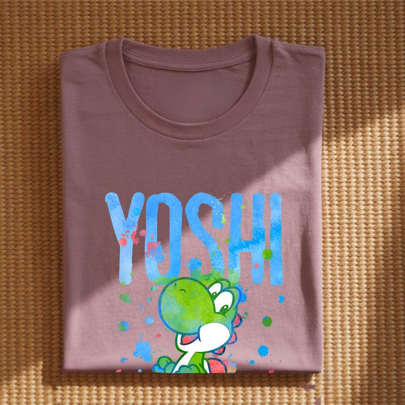May include: A mauve t-shirt with the word "YOSHI" in blue, and a green cartoon character. The character has a green head and body, a white belly and red accents. Colourful paint splatters surround the character and text.