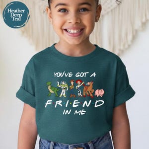May include: Teal short-sleeve t-shirt featuring the phrase "YOU'VE GOT A FRIEND IN ME" with cartoon characters. The design includes a green dinosaur, a space ranger, Woody, Jessie, a horse, and a pig. The shirt is worn by a child.
