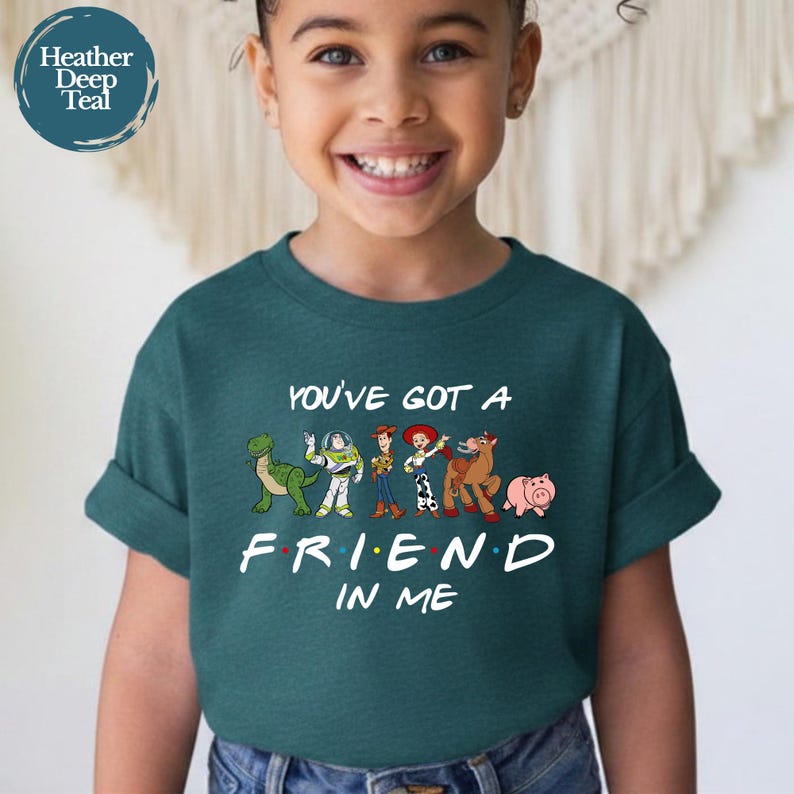 May include: Teal short-sleeve t-shirt featuring the phrase "YOU'VE GOT A FRIEND IN ME" with cartoon characters. The design includes a green dinosaur, a space ranger, Woody, Jessie, a horse, and a pig. The shirt is worn by a child.