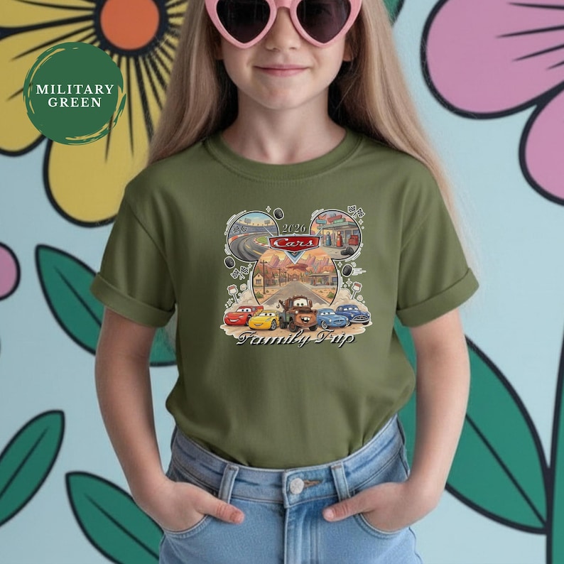 Puede incluir: Camiseta verde oliva con un gr&aacute;fico de los personajes de Cars y el texto "2020 Cars Family Trip". El dise&ntilde;o incluye im&aacute;genes de coches de carreras, una carretera y una gasolinera. La camiseta la lleva una persona.