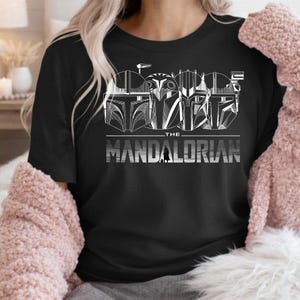 May include: Black t-shirt featuring a white graphic of four Mandalorian helmets and the text "THE MANDALORIAN" in a bold, silver font. The design includes a small silhouette of a Mandalorian figure. The shirt is a crew neck.