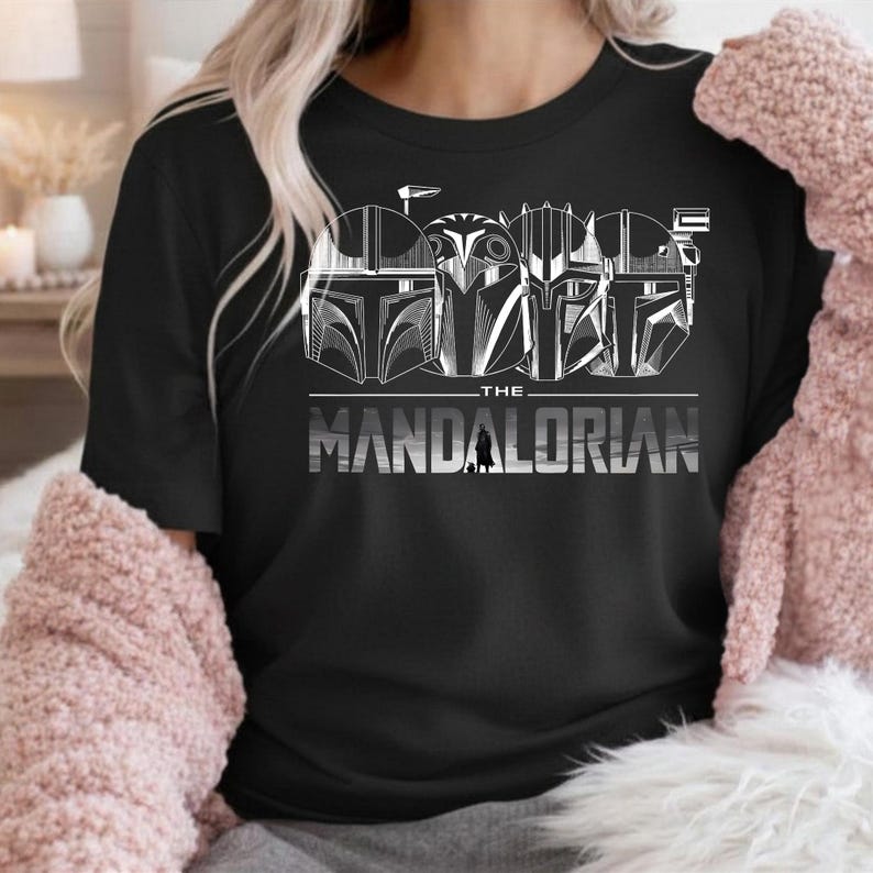 May include: Black t-shirt featuring a white graphic of four Mandalorian helmets and the text "THE MANDALORIAN" in a bold, silver font. The design includes a small silhouette of a Mandalorian figure. The shirt is a crew neck.