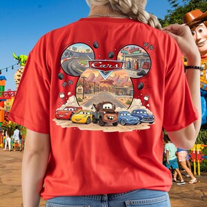 Pu&ograve; includere: Una t-shirt rossa con una stampa grafica del logo Disney Pixar Cars e personaggi tra cui Saetta McQueen, Cricchetto e altri veicoli. Il design ha la forma di una silhouette di Topolino con scene di una pista da corsa e una citt&agrave;.