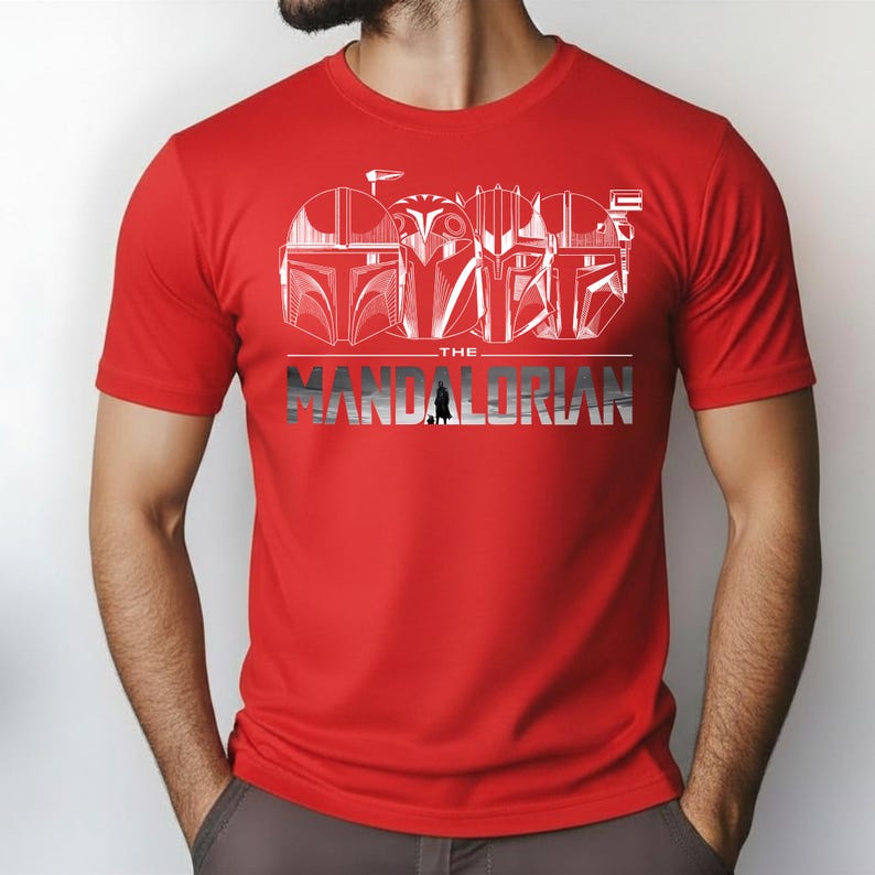 May include: Red t-shirt featuring a white graphic of Mandalorian helmets and the text "THE MANDALORIAN" in silver and black. The design includes a silhouette of the Mandalorian. Short-sleeved, crew neck.