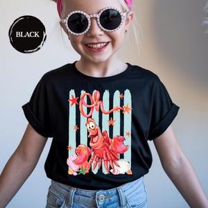 Pu&ograve; includere: Una t-shirt nera a maniche corte con una stampa grafica di un granchio rosso e la parola Oh in corsivo rosso. Il design include strisce verticali azzurre, piccole stelle marine arancioni e varie conchiglie nella parte inferiore del disegno.
