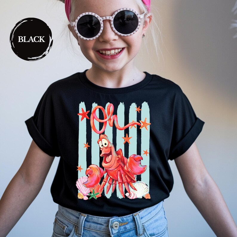 Pu&ograve; includere: Una t-shirt nera a maniche corte con una stampa grafica di un granchio rosso e la parola Oh in corsivo rosso. Il design include strisce verticali azzurre, piccole stelle marine arancioni e varie conchiglie nella parte inferiore del disegno.