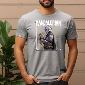 May include: Gray t-shirt featuring a graphic of The Mandalorian and Grogu. The design includes the text "MANDALORIAN - THIS IS THE WAY" in a white rectangular frame. The Mandalorian is depicted in armor, holding Grogu.