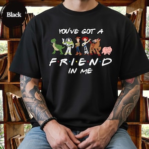 May include: Black t-shirt featuring the phrase "You've Got A Friend In Me" with cartoon characters. The design includes a green dinosaur, Buzz Lightyear, Woody, Jessie, a horse, and a pig. The shirt is a crew neck.