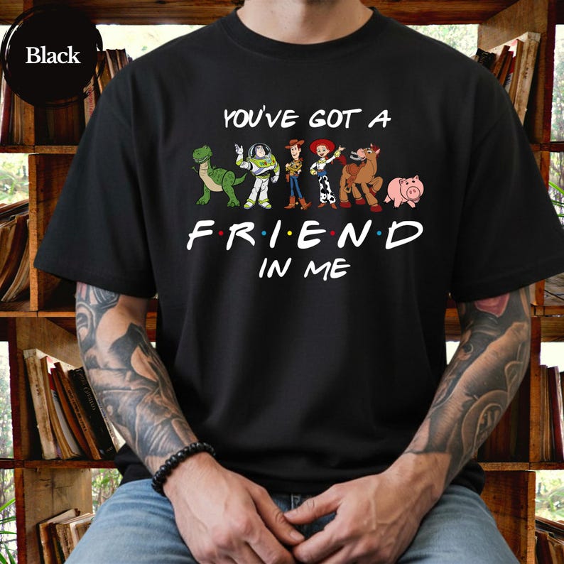 May include: Black t-shirt featuring the phrase "You've Got A Friend In Me" with cartoon characters. The design includes a green dinosaur, Buzz Lightyear, Woody, Jessie, a horse, and a pig. The shirt is a crew neck.