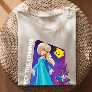 May include: Cream-colored t-shirt featuring a graphic of a princess in a blue dress with a crown, a yellow star, and a space background. The text "PROTECT THE COSMOS" is printed vertically on the left side of the graphic.