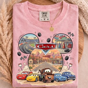 May include: A light pink Comfort Colors t-shirt featuring a Cars graphic. The graphic includes the Cars logo, race track, gas station, and cartoon cars. The shirt has a crew neck and short sleeves. A gold necklace with a sun pendant is visible.