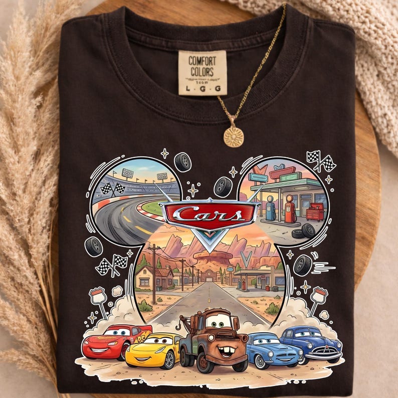 May include: Dark brown t-shirt featuring a colorful graphic from the movie "Cars." The design includes the word "Cars" in a red and white logo, race cars, a racetrack, and a desert town scene. The shirt has a Comfort Colors tag.