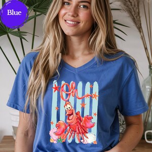 Pu&ograve; includere: Una t-shirt blu con scollo a V caratterizzata da una stampa grafica di un granchio rosso con il testo "Oh" sopra. Il design include pennellate verticali azzurre, stelle marine arancioni e varie conchiglie nei colori rosa, bianco e beige.