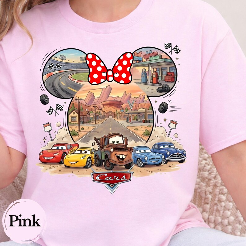 Pode incluir: Uma t-shirt rosa claro com um gr&aacute;fico das personagens do filme Carros da Disney, incluindo Rel&acirc;mpago McQueen e Mate. O design tem a forma de uma cabe&ccedil;a do Mickey com um la&ccedil;o vermelho de bolinhas brancas, mostrando uma estrada no deserto.