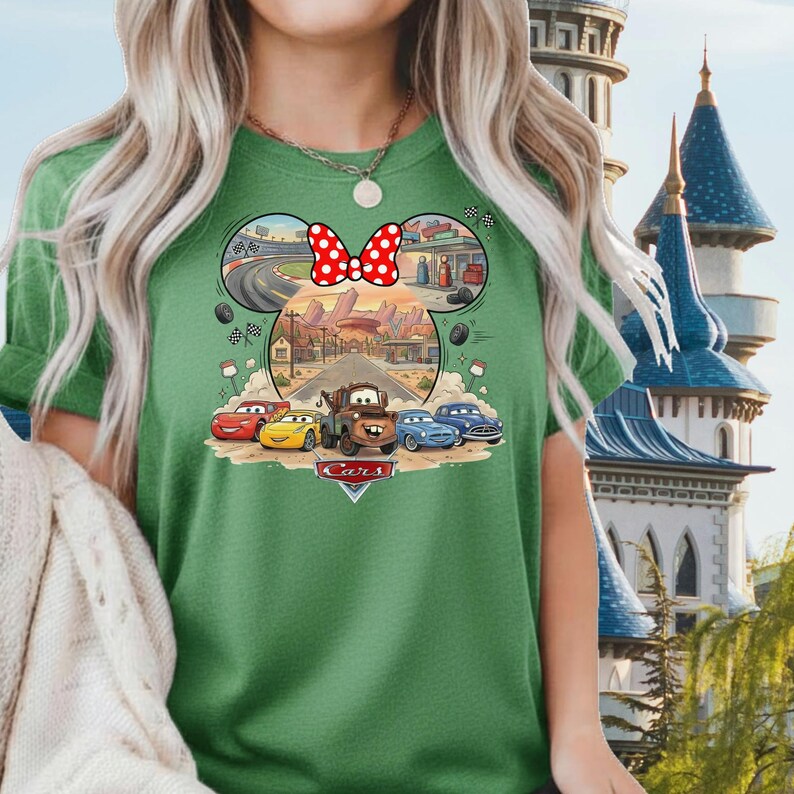 Pode incluir: Uma t-shirt verde com um gr&aacute;fico das personagens do filme Carros da Disney, incluindo Rel&acirc;mpago McQueen e Mate, dentro de uma moldura em forma de orelhas do Mickey Mouse. O design inclui um la&ccedil;o vermelho &agrave;s bolinhas no topo.